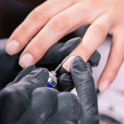 Close-up professional beautician hands working with electric drill on client fingernails. Procedure applying artificial fingernails. Hands of manicurist with tool. Manicurist in gloves removes cuticle